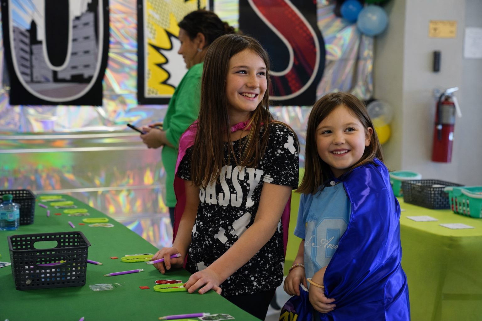 Two smiling girls wearing superhero capes at a craft table